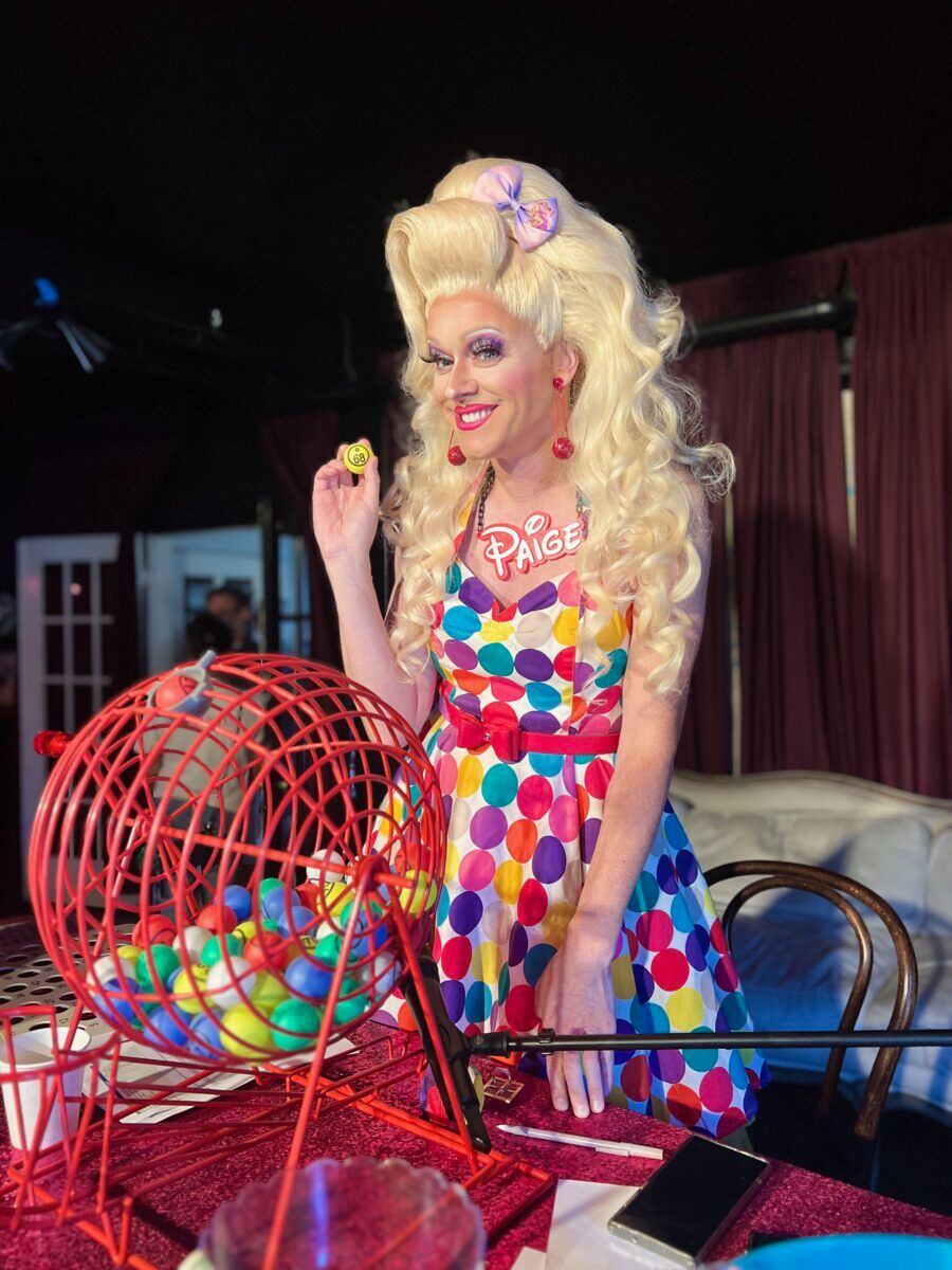 An adorable bubblegum queen holds a bingo ball while smiling at the audience out of frame
