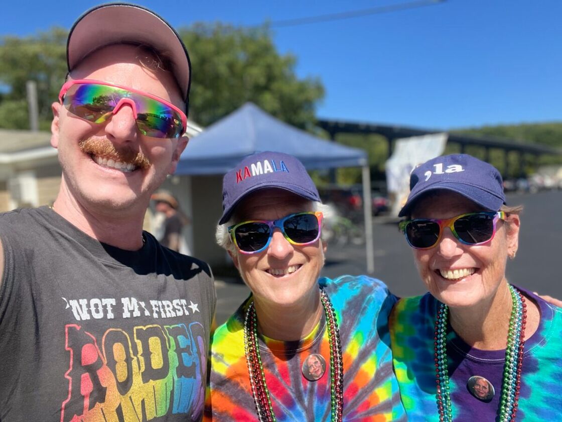 Three folks stand smiling for a selfie. They are sporting colorful beads and tye-dye shirts