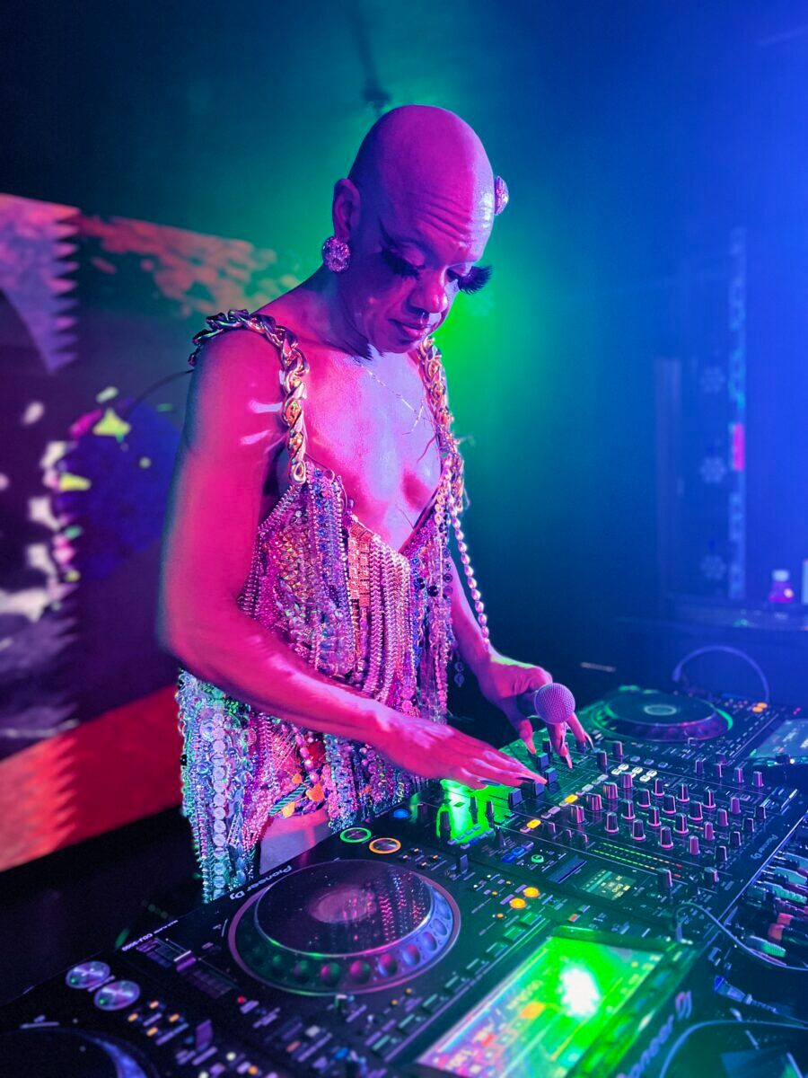 A bald drag queen stands in front of a DJ set up, focused on the knobs