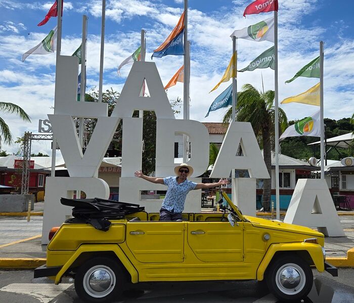 The author stands in the back of a yellow convertible in front of a giant sculpture of the words "La Vida Bella"