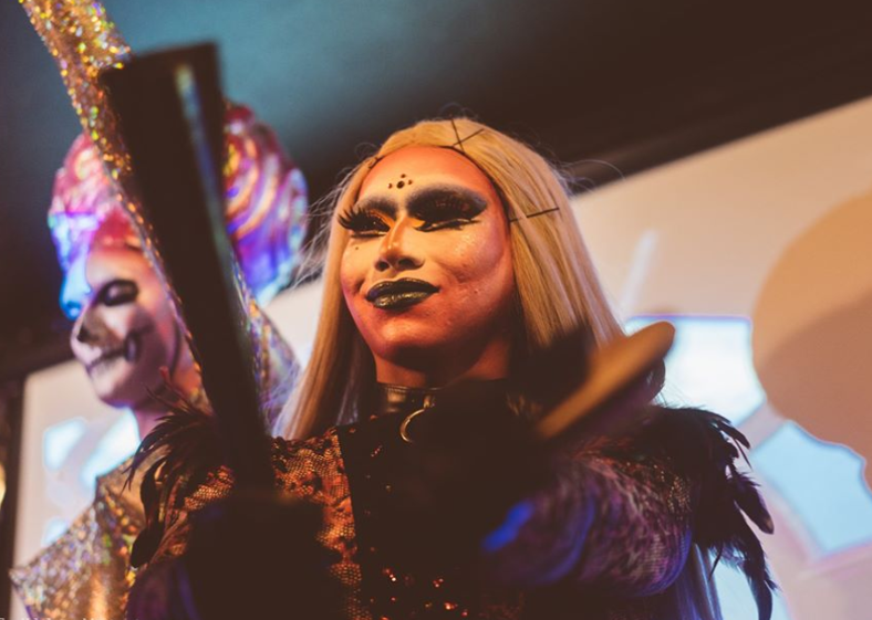 A smiling drag queen wearing feathers and futuristic makeup smiles from the stage.