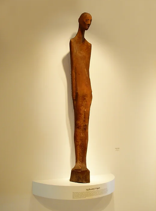 A striking statue carved from wood depicting a thin and gaunt figure with a haunting face. 