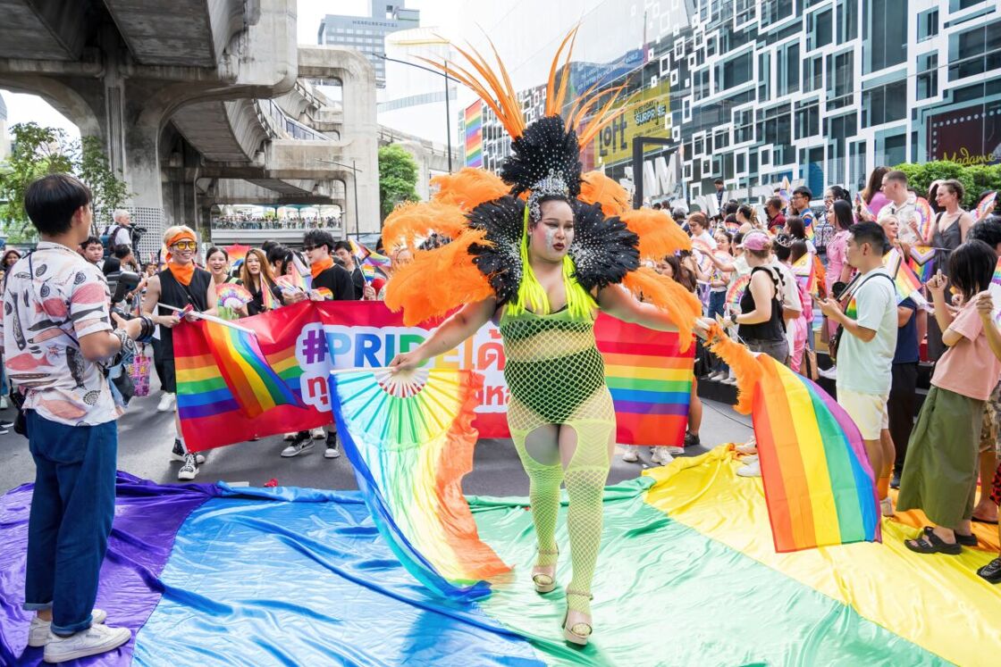 A drag performer in a vibrant feathered costume, featuring orange and black plumes and a neon green outfit, dancing at a Pride parade. Behind them, a group of people with rainbow flags and a large banner that reads “#PRIDE”