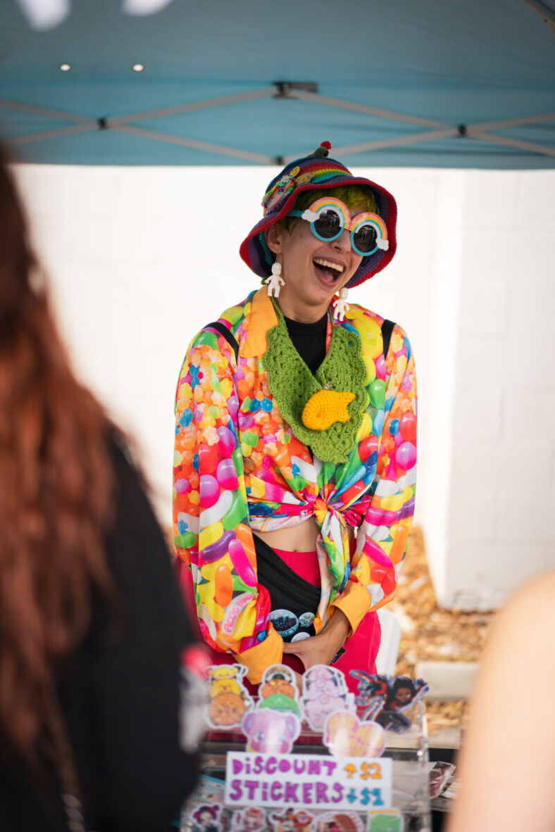 A colorfully dressed person smiles broadly as she stands behind her merchandise. She wears rainbow sunglasses, a long-sleeved jelly bean-print shirt and a rainbow bucket hat. Photographer: Tyler Karon