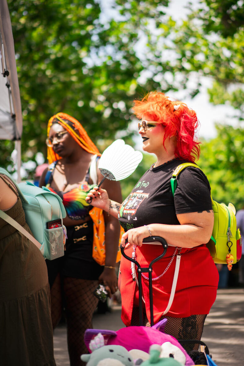 One person stands with a small cart full of Squishmallows and holds a small fan in her hand. Her hair is dyed bright orange and red and she is wearing a bright red skirt and fishnet tights. Photographer: Jordan Macaulay