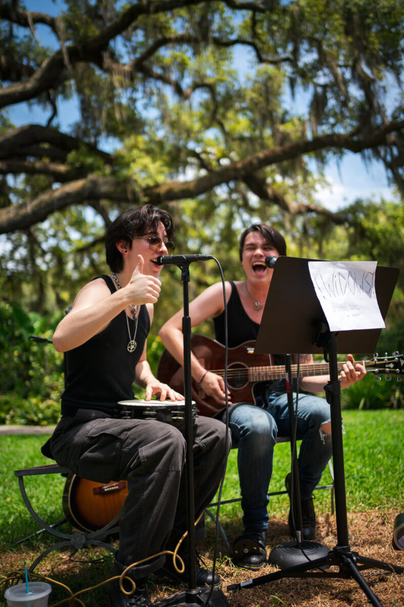 Two young musicians sit in front of microphones, smiling. One holds a guitar, the other a tambourine. Both wear black tank tops and have black hair. Photographer: Tyler Karon