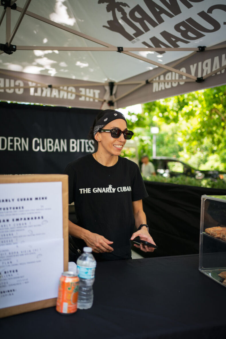 A smiling server stands under a tent wearing a black shirt with the words "The gnarly Cuban" on it. They are also wearing sunglasses and a black bandana. Photographer: Tyler Karon