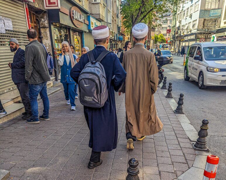 Two traditionally dressed men walking down a street in Fatih. Men in drab outfits walk on the sidewalk