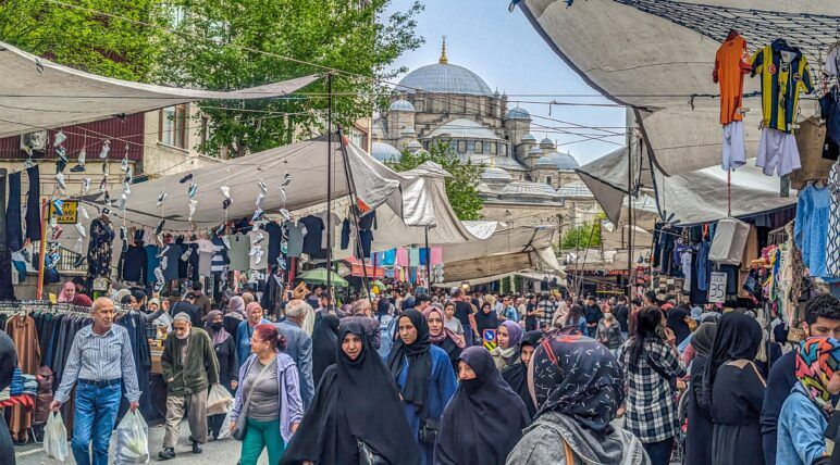 Street packed with shoppers, including men dressed in typical Western clothing, while most women are dressed traditionally. Street view of Fatih's crowded outdoor market with a mosque in the background
