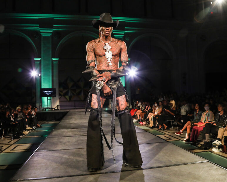 Tattooed Black man in a black cowboy hat, skirt, and chaps with a huge silver necklace hanging down his chest