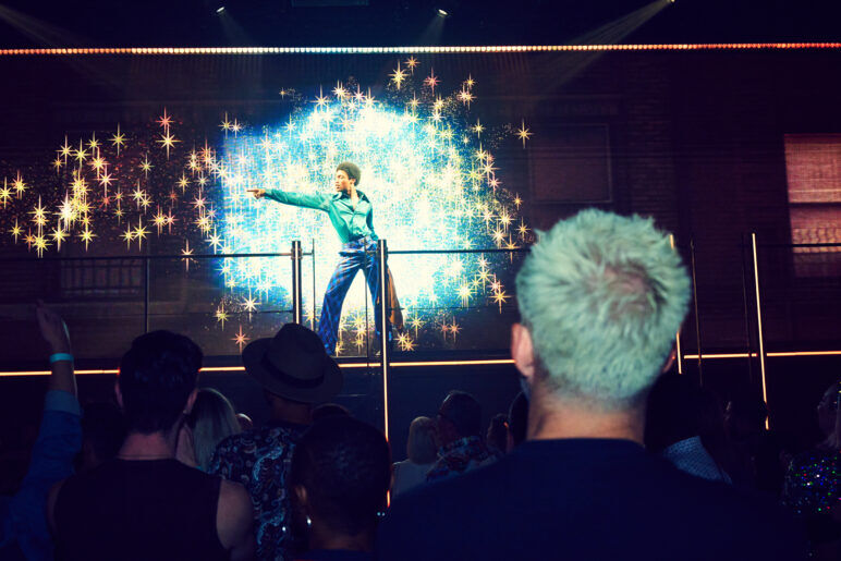 A young Black man points in a classic disco pose on stage at DISCOSHOW as the crowd looks on