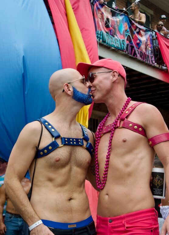 Two shirtless men, share an adorable kiss. One is wearing a pink harness, while the other one has a blue harness and sparkly blue beard