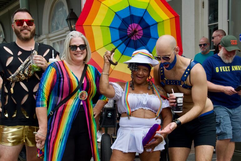 Four friends smile while holding a rainbow unbrella. They are all having fun. One has a glittery blue beard and blue harness on