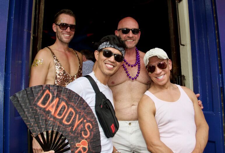 Four men smile at the camera. One holds a big fan that has the word 