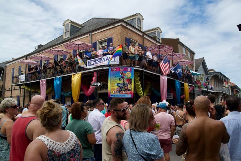 A packed street with rainbow flags and a New Orleans style building with an equally packed balcony