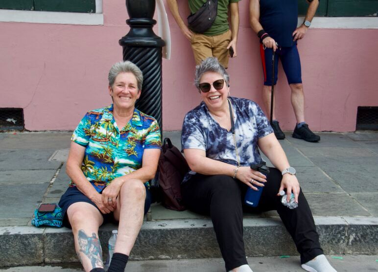 Two short-haired women take a break sitting on the curb of the street. They are smiling broadly and drinking water