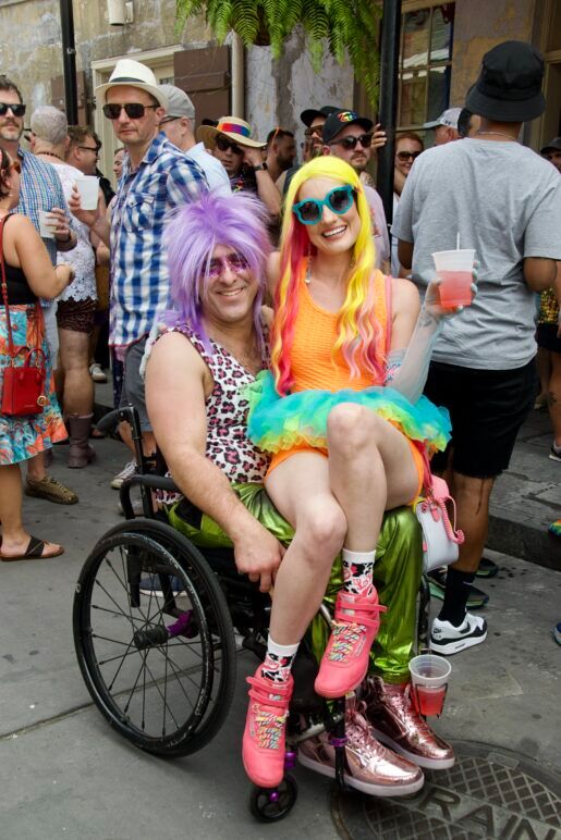 A woman sits daintily on the lap of a man in a wheel chair. They are both wearing colorful wigs and bright clothing, smiling at the camera.