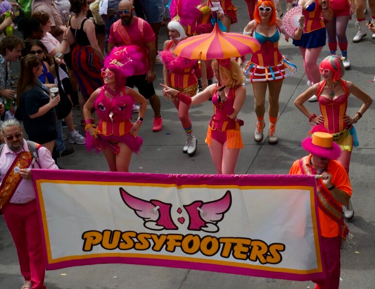 A group of parade walkers all wear bright and funky costumes while holding a banner that reads "Pussyfooters"