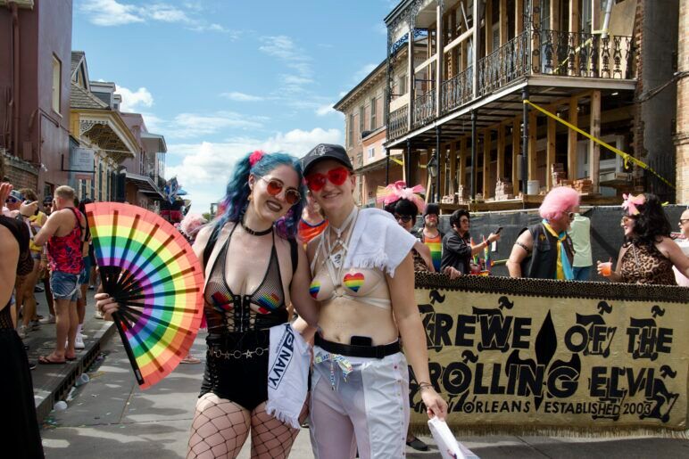 Two femme presenting people with rainbow heart stickers on their chests smile at the camera while standing on the street