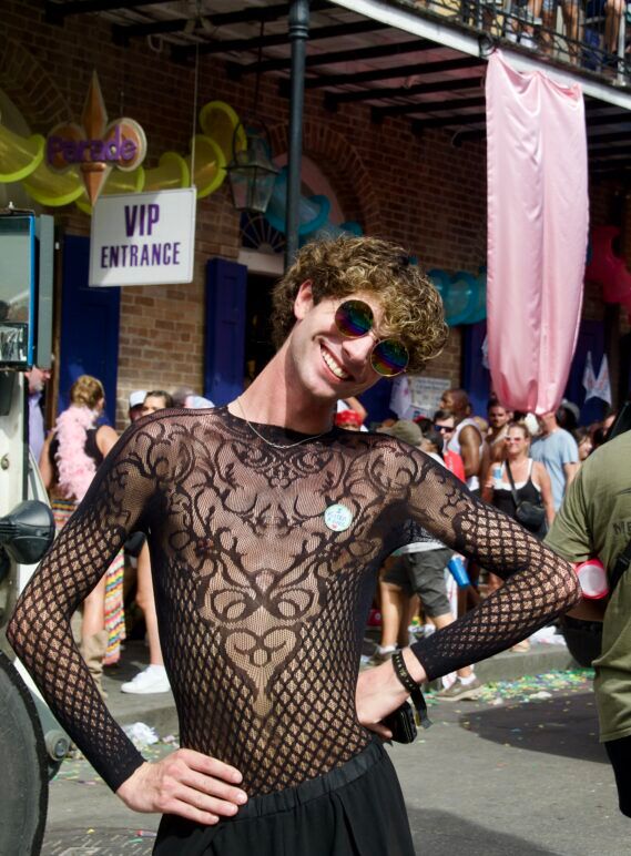 A cutie with curly hair and sunglasses poses and smiles at the camera while wearing a Victorian print mesh shirt