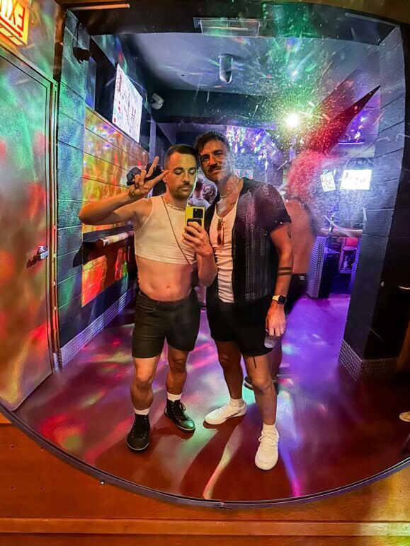 A mirror selfie of Ian and Smith at a bar with colorful, disco-like lighting reflecting off the walls. Ian, on the left, is wearing a white crop top, shorts, and sneakers, posing with a peace sign. Smith, on the right, is dressed in a black shirt over a white tank top, shorts, and white sneakers. The background is filled with partygoers, creating a fun and lively atmosphere.