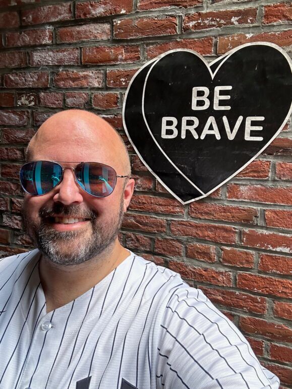 JJ Keyes next to a brick wall with graffiti that says "Be Brave"