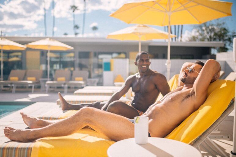 Two men in swimsuits sit poolside in beach chairs