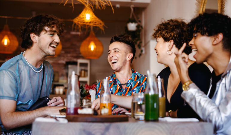 A group of friends laughing and enjoying drinks together at an LGBTQ+ inclusive restaurant. The atmosphere is warm and inviting, with soft lighting from hanging lanterns, highlighting the joy and connection among the group.