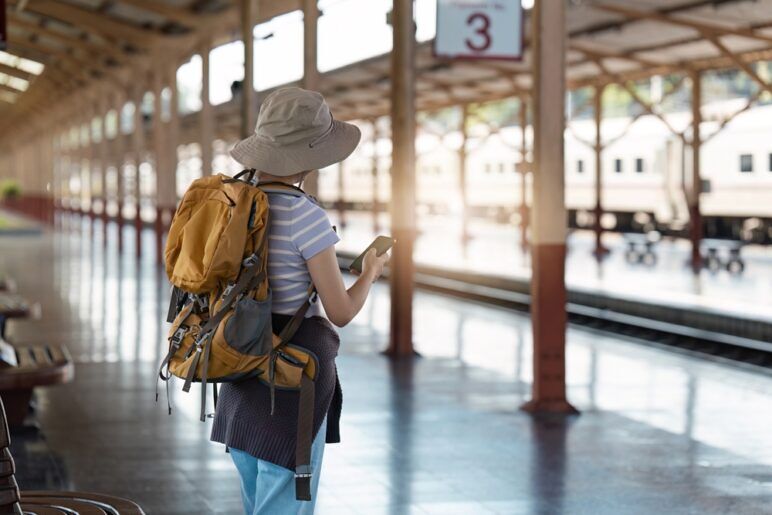 Solo Traveler with Backpack and Hat Waiting at Train Station Platform, Checking Phone for Travel Updates