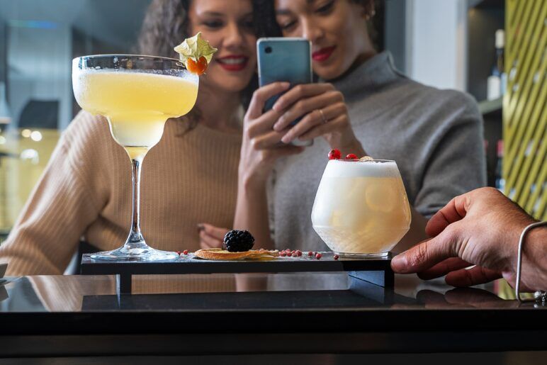 Two women, smiling and capturing a photo of elegant cocktails with their phone at an LGBTQ+-friendly bar. The drinks feature artistic garnishes, while one person’s hand is adjusting a glass, highlighting the communal experience of enjoying drinks together.