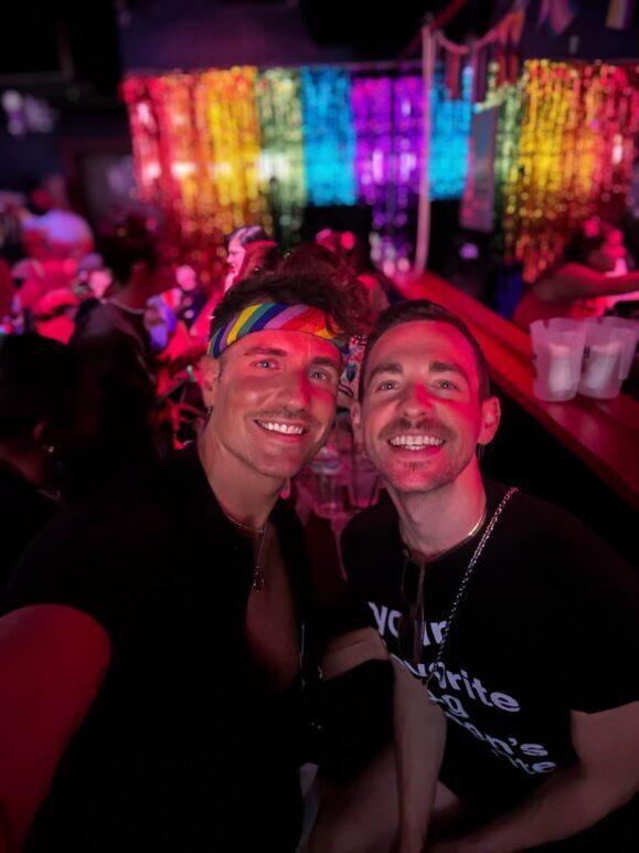 A selfie of two smiling gay men at a lively bar with a rainbow backdrop made of shimmering tinsel. The man on the left is wearing a colorful rainbow headband and a black shirt, while the man on the right wears a black T-shirt with white text and a silver chain. 