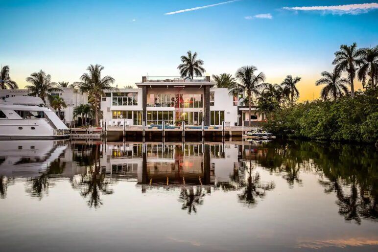 The sun sents behind a villa situated on the ocean with a yacht tied up out front