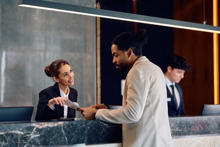 A man checks in at a hotel front desk, engaging with a friendly receptionist who is handing him his room key and paperwork. Both are professionally dressed, with the scene highlighting a welcoming atmosphere at a high-end hotel. Another staff member is visible in the background, attending to a separate task.