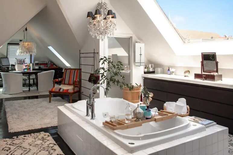 A marble tub sits next to a skylight with a chairs surround a table lit by a giant chandelier in the background