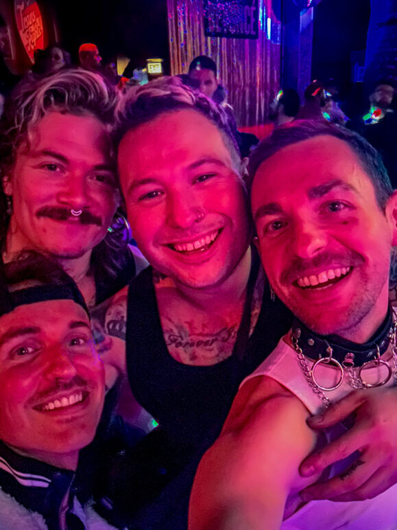 A vibrant group selfie taken at a bar with colorful red and purple lighting. Four smiling men are closely gathered, with the person on the right wearing a collar and chain accessory, and the others sporting a variety of piercings and tattoos. The background is filled with more partygoers, and the atmosphere is lively and energetic, with hints of glitter and neon adding to the party vibe.