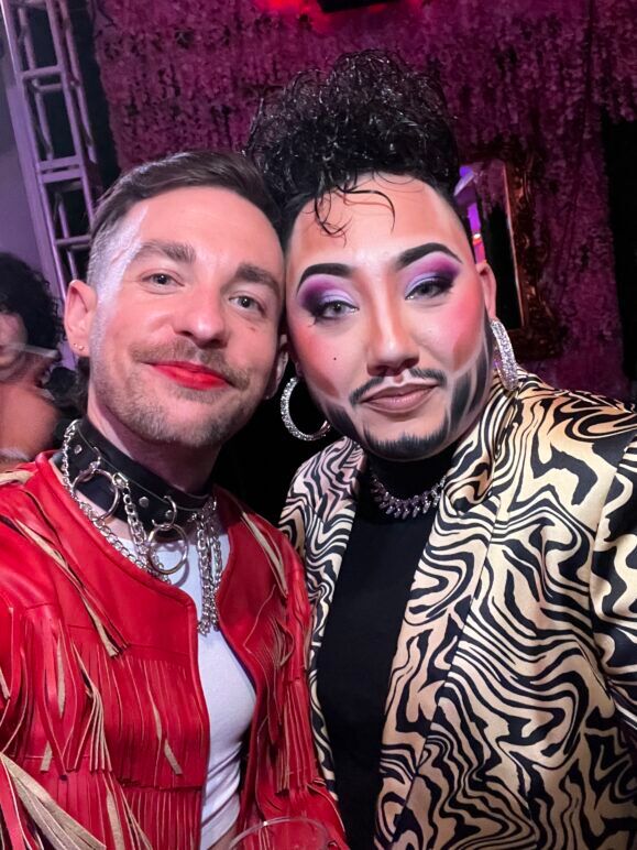 A selfie of Ian and drag king Tenderoni at a lively event. Ian, smiling brightly, is wearing a black shirt and chain, while Tenderoni, dressed in a shiny, zebra-esque 80's suit, strikes a playful pose. The background is filled with people, vibrant lighting, and an upbeat atmosphere, capturing the fun and celebratory energy of the event.