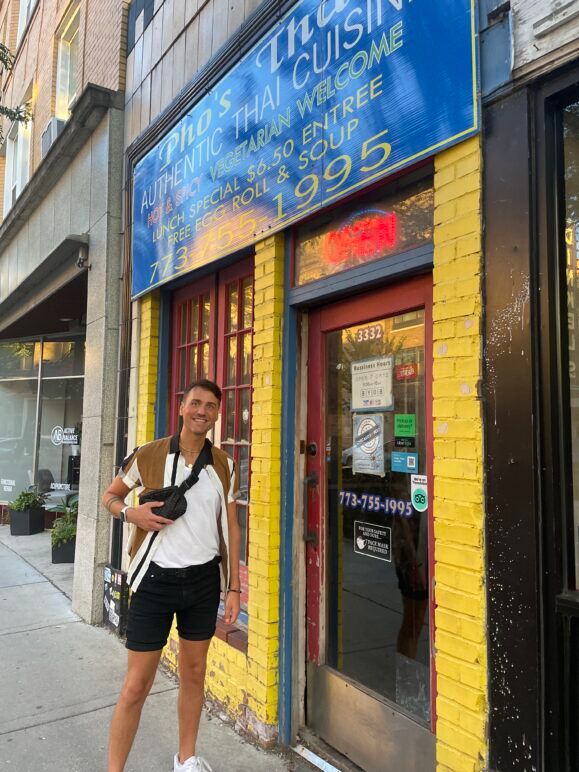 Smith stands in front of Pho’s Thai Cuisine, smiling warmly at the camera. Wearing a casual outfit with a crossbody bag, they pose outside the restaurant, which has bright windows and signage visible in the background. The setting captures a relaxed moment before enjoying a meal at the cozy, local Thai spot.
