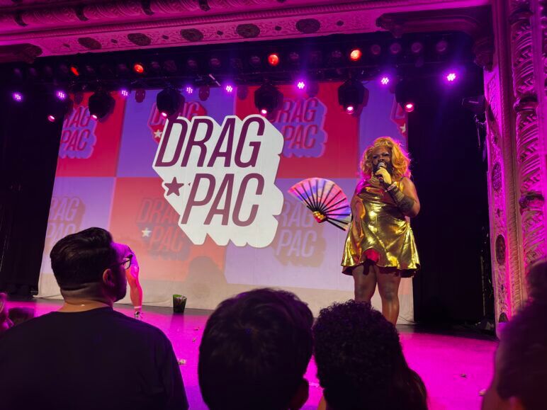 Alt text:

Drag performer Lucy Stoole stands on stage in a gold dress, holding a fan, and speaking into a microphone during a Drag PAC event. The backdrop displays the Drag PAC logo in large letters, while the audience watches her performance. The venue is illuminated with purple stage lights, and the ornate architecture of the stage adds to the lively, political drag atmosphere.