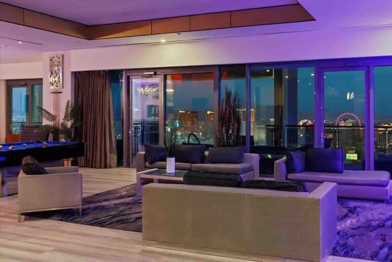View of the penthouse living room, lit with purple light, with a view of the Las Vegas skyline through the floor-to-ceiling windows