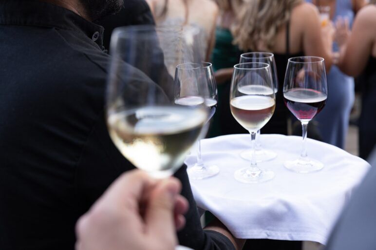 A close-up of a tray with several glasses of wine being served at a hotel bar, with a focus on a person holding a glass of white wine in the foreground.