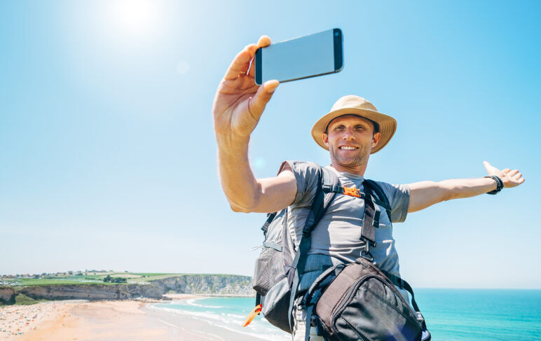 A backpacker traveling alone on the beach taking a selfie wearing a hat. 