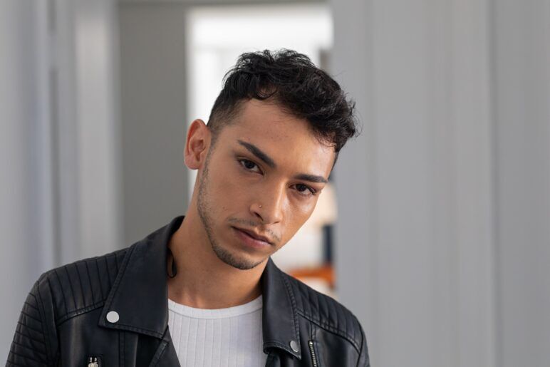 A young biracial man with short curly hair and a nose piercing, wearing a black leather jacket and a white ribbed shirt, gazing directly into the camera with a confident expression.