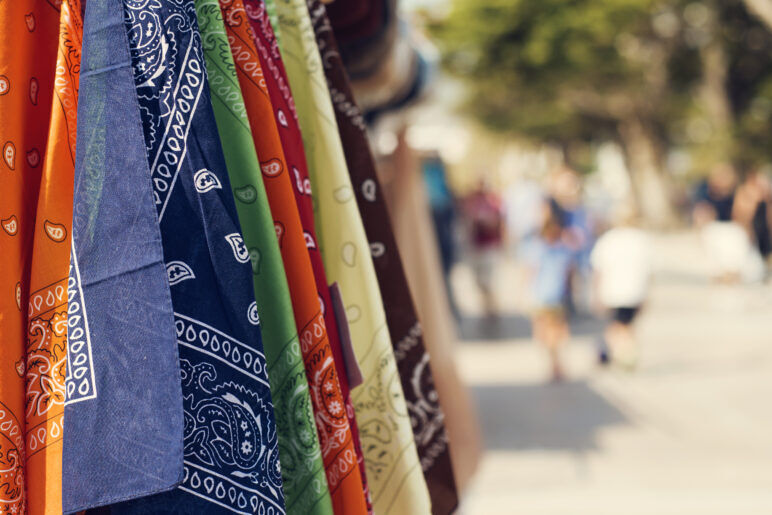 A close-up of various colored hankies, including orange, navy blue, green, red, yellow, and brown, hanging in a row with a blurred outdoor setting and people walking in the background.