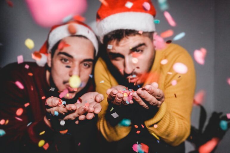 Two men wearing Santa hats blow colorful confetti toward the camera, celebrating the holiday season with joy and festive cheer.