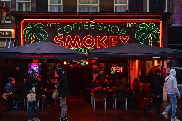 The exterior of Smokey Coffeeshop in Amsterdam at night, featuring a vibrant neon sign with green palm trees and red text that reads “Coffeeshop Smokey.” People are gathered under black awnings that also display the coffeeshop’s name