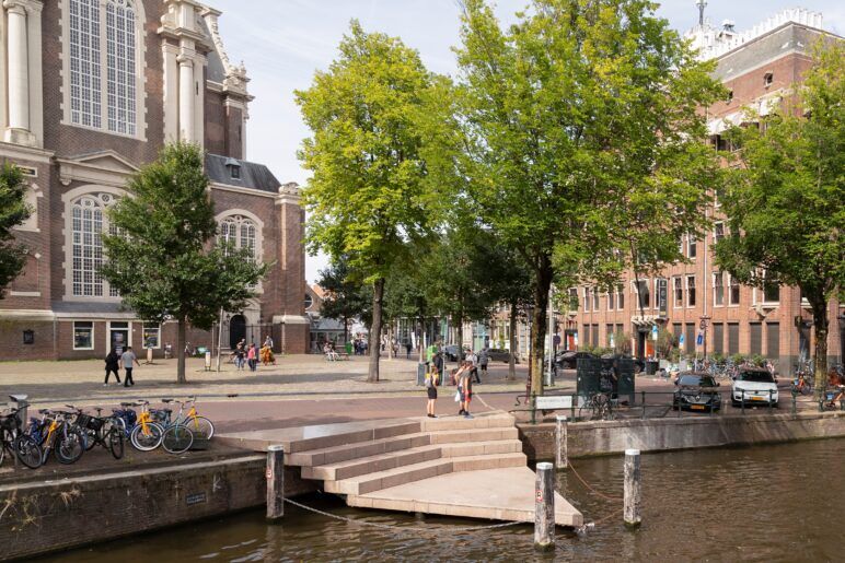 The image shows a picturesque canal-side scene in Amsterdam including the Homomonument. In the foreground, a set of steps leads down to the canal, bordered by wooden posts and a small docking area. The background features a historical brick building with large, ornate windows. People are strolling and enjoying the area, and bicycles are parked nearby.