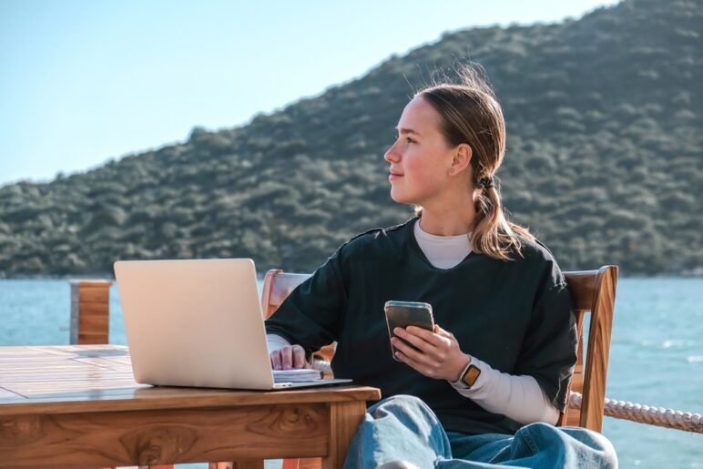 A woman works on a laptop outside with a mountain behind her