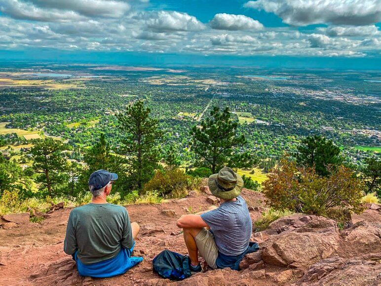 Two men sit on a bluff overlooking a green valley with their backs to the camera.