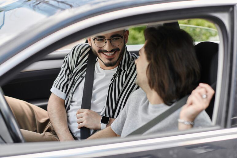 A gay couple smiles and laughs while getting buckled up in a car before heading out on a staycation nearby.