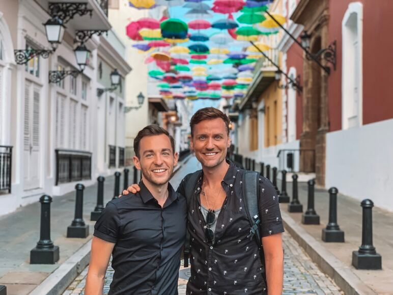 Ian and their partner Smith smiling and standing close together on a colorful street under an umbrella canopy in San Juan, Puerto Rico.